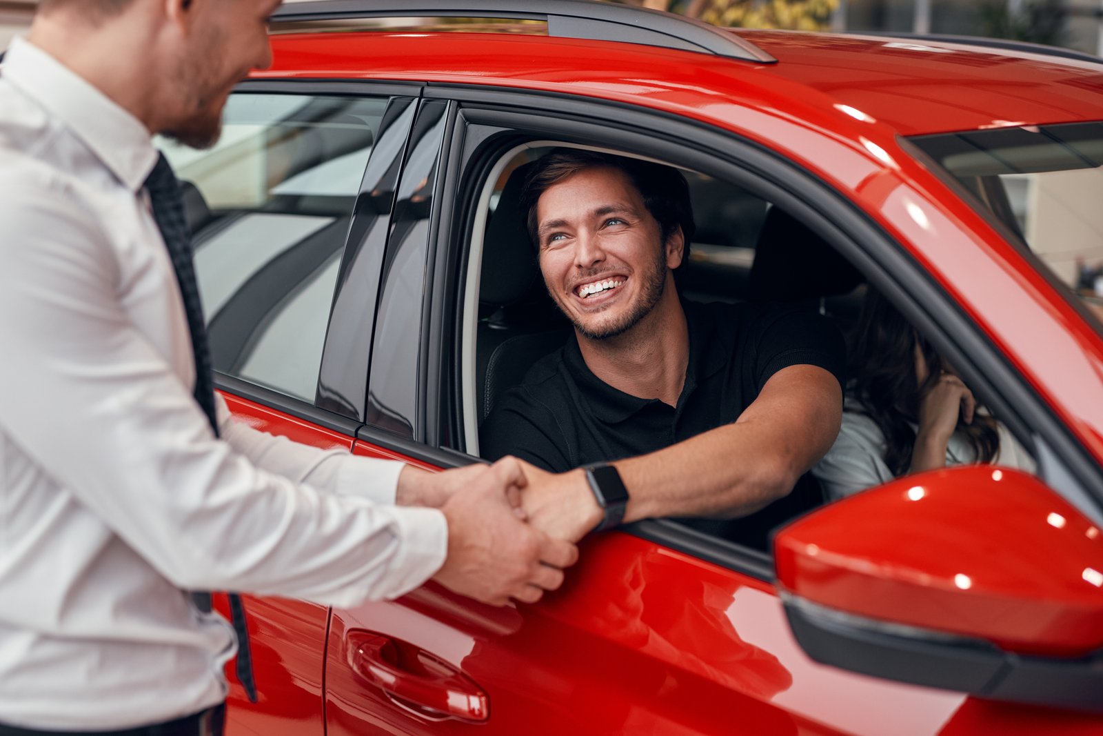 Cheerful male customer smiling and shaking hand of manager while sitting inside new red vehicle and sealing deal in car dealership